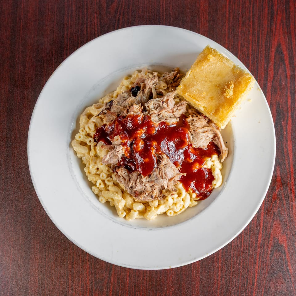 Plate of creamy macaroni topped with pulled pork, barbecue sauce, and cornbread on a wooden table.