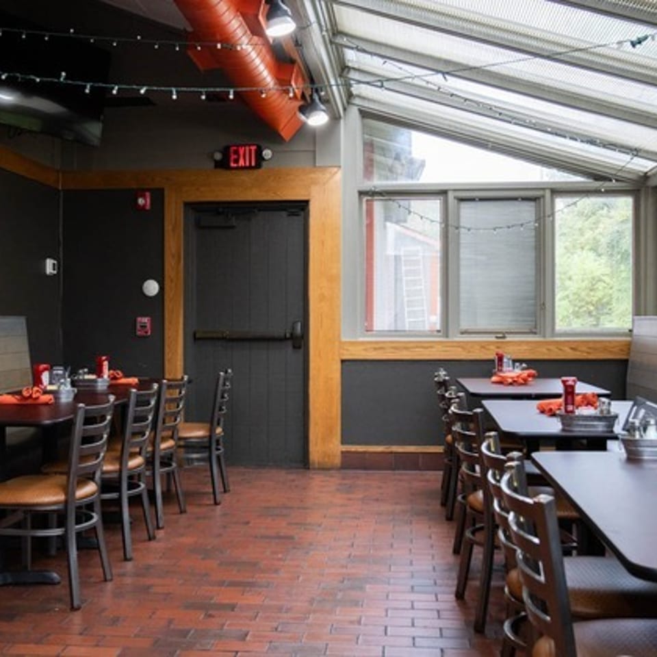 Bright enclosed restaurant dining area with booth seating, wooden tables, orange napkins, string lights, and large windows.