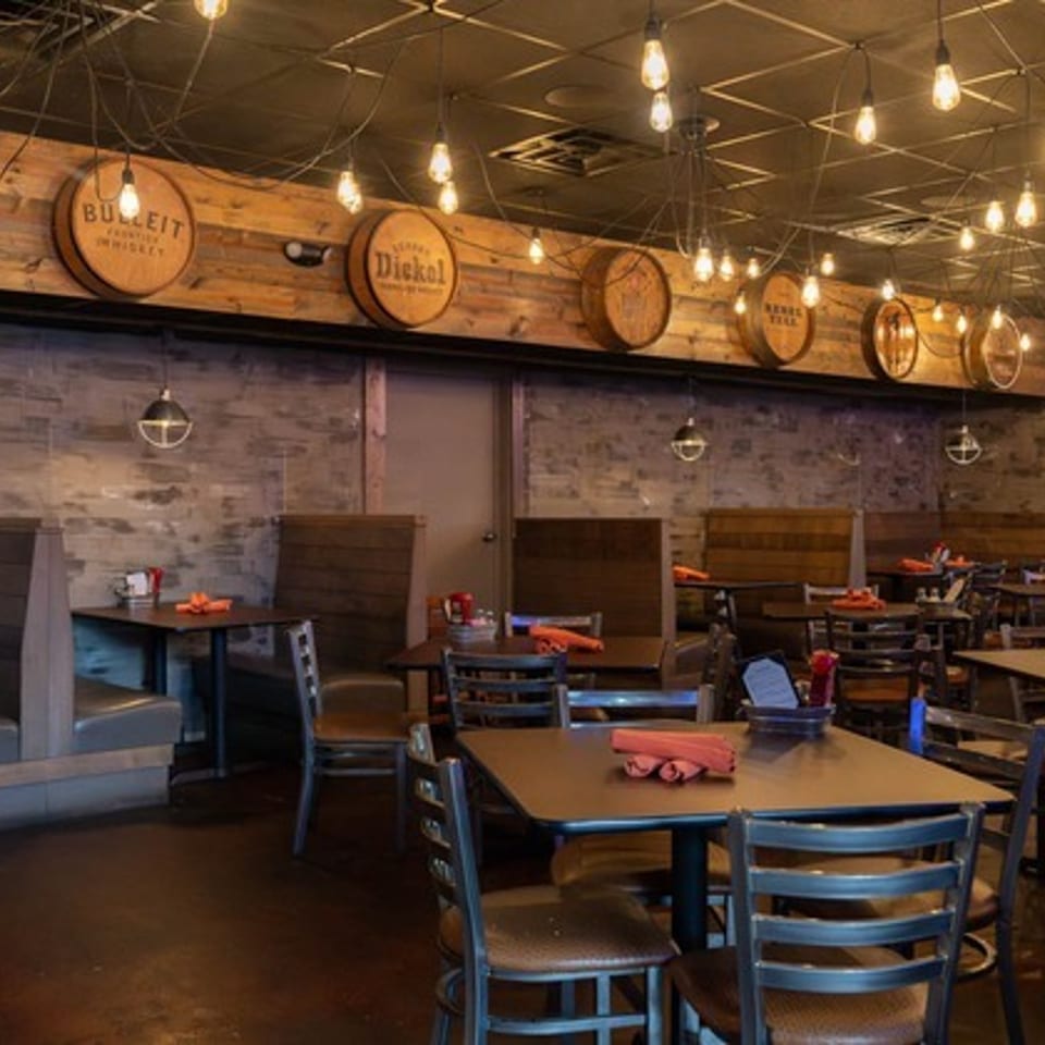 Rustic restaurant dining room with wooden tables, booths, exposed brick walls, hanging lights, and beer barrel decorations.