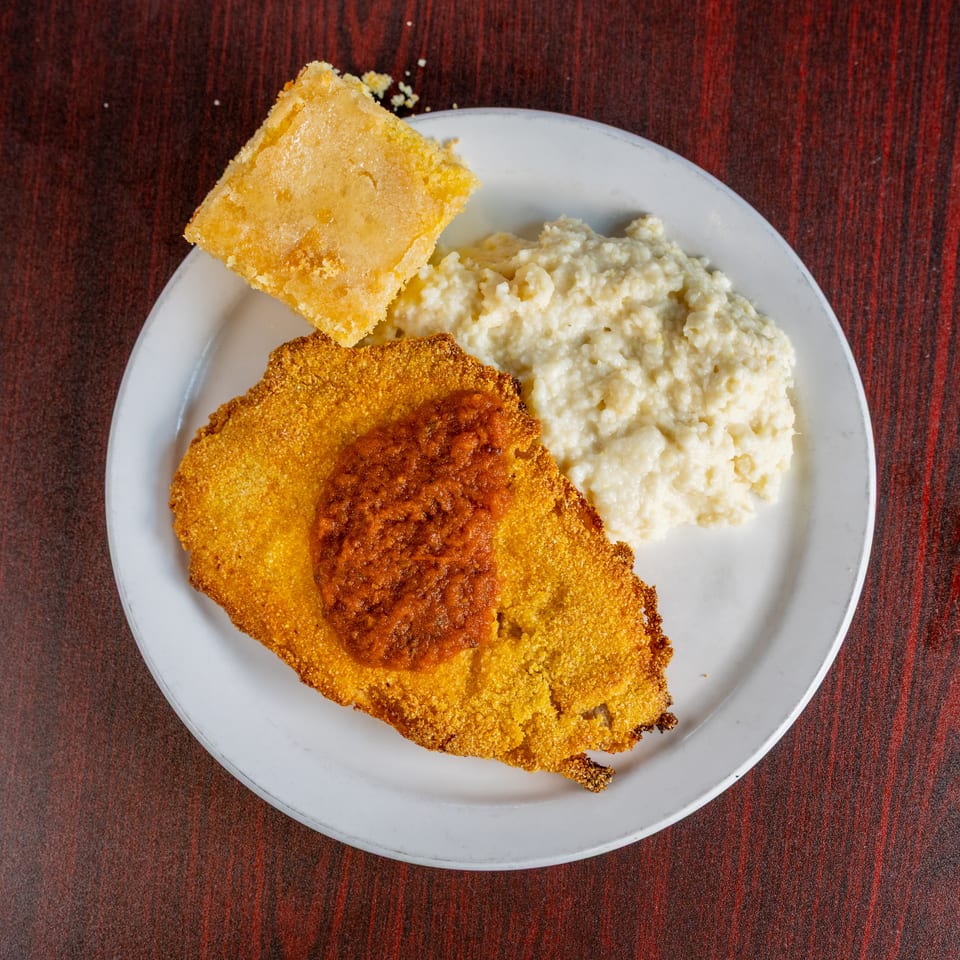 Plate of breaded fried fish, creamy mashed potatoes, and cornbread on a dark wood table.