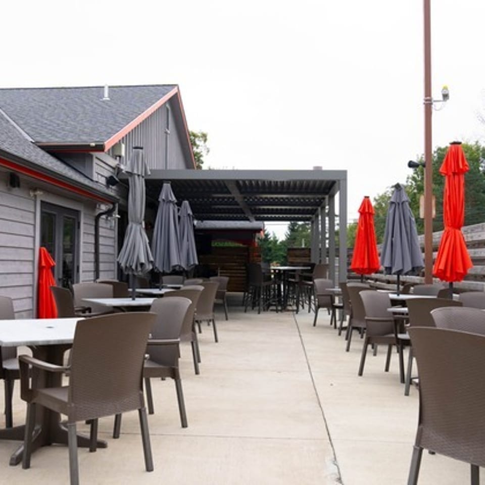 Outdoor restaurant patio with tables, chairs, red and gray umbrellas, wooden privacy fencing, and a covered dining area.