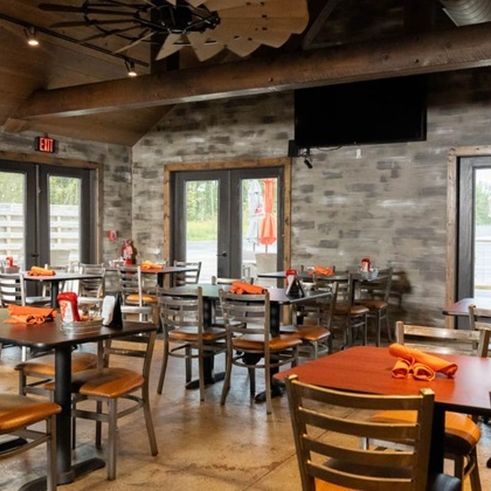 Rustic restaurant dining room with wood-paneled walls, exposed ductwork, neatly arranged tables, and orange napkins.