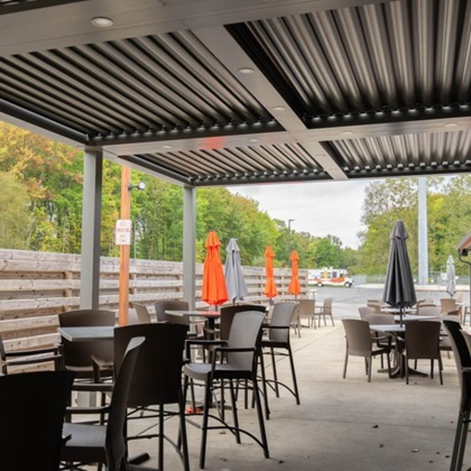 Covered outdoor restaurant patio with tables, chairs, colorful umbrellas, wooden fencing, and trees beyond.