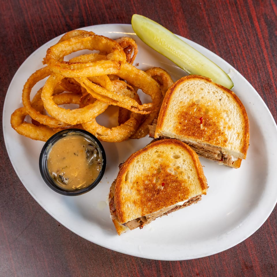 Grilled sandwich served with crispy onion rings, dipping sauce, and a pickle spear on a white plate.