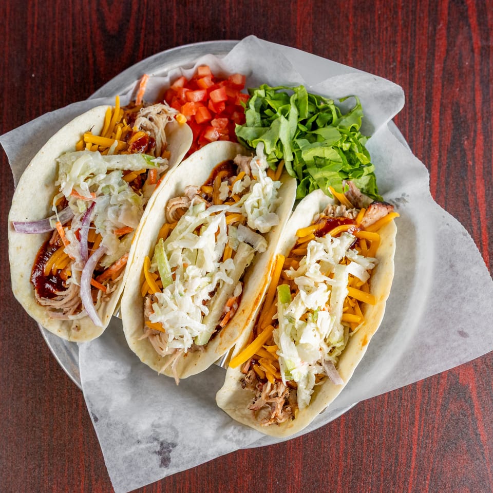 Three soft tacos filled with shredded meat, cheddar cheese, creamy slaw, lettuce, and diced tomatoes on a paper-lined plate.