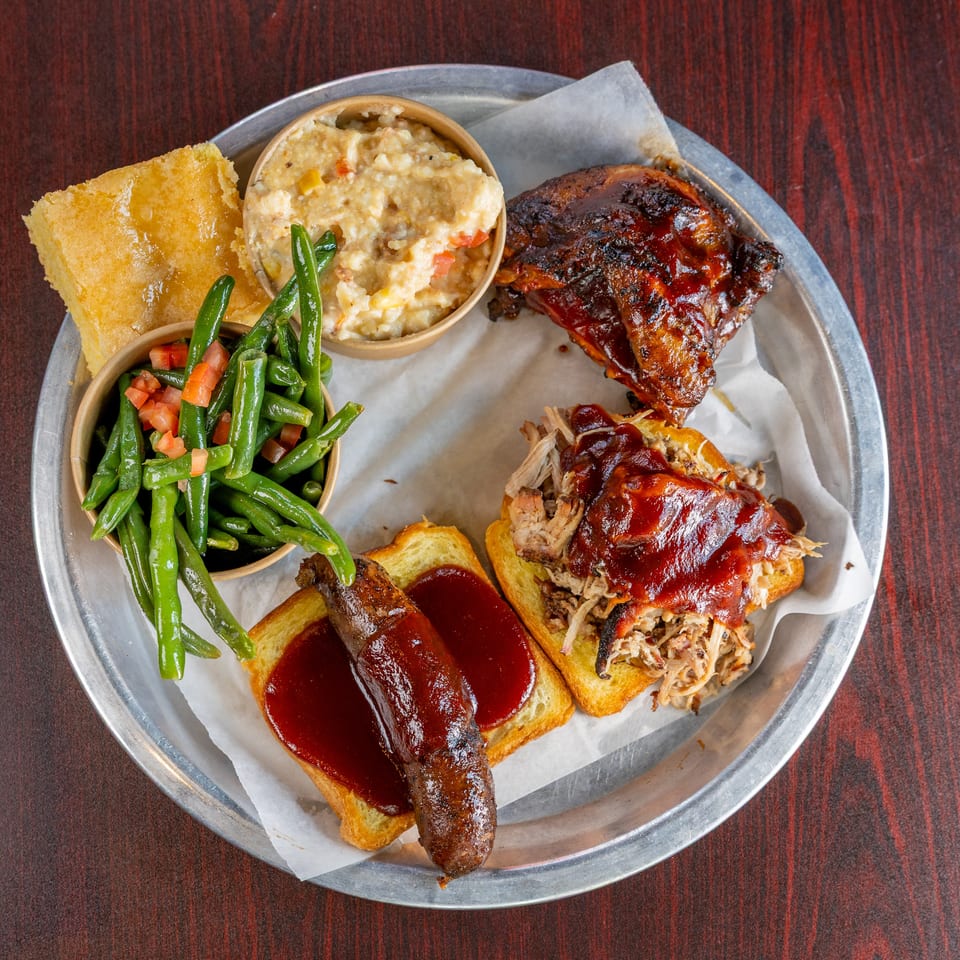Barbecue platter with sauced ribs, pulled pork on toast, green beans, creamy side dish, and cornbread on a metal tray.